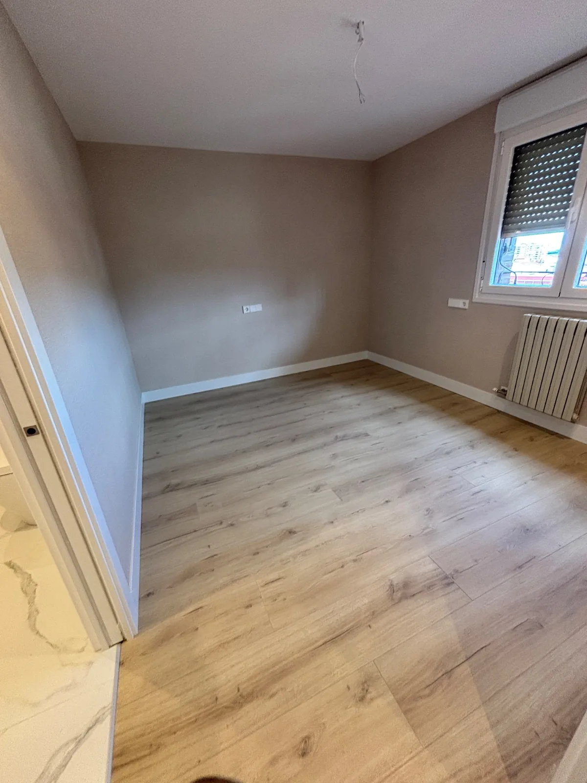 Dormitorio vacío recién reformado con suelo laminado, radiador y ventana con persiana