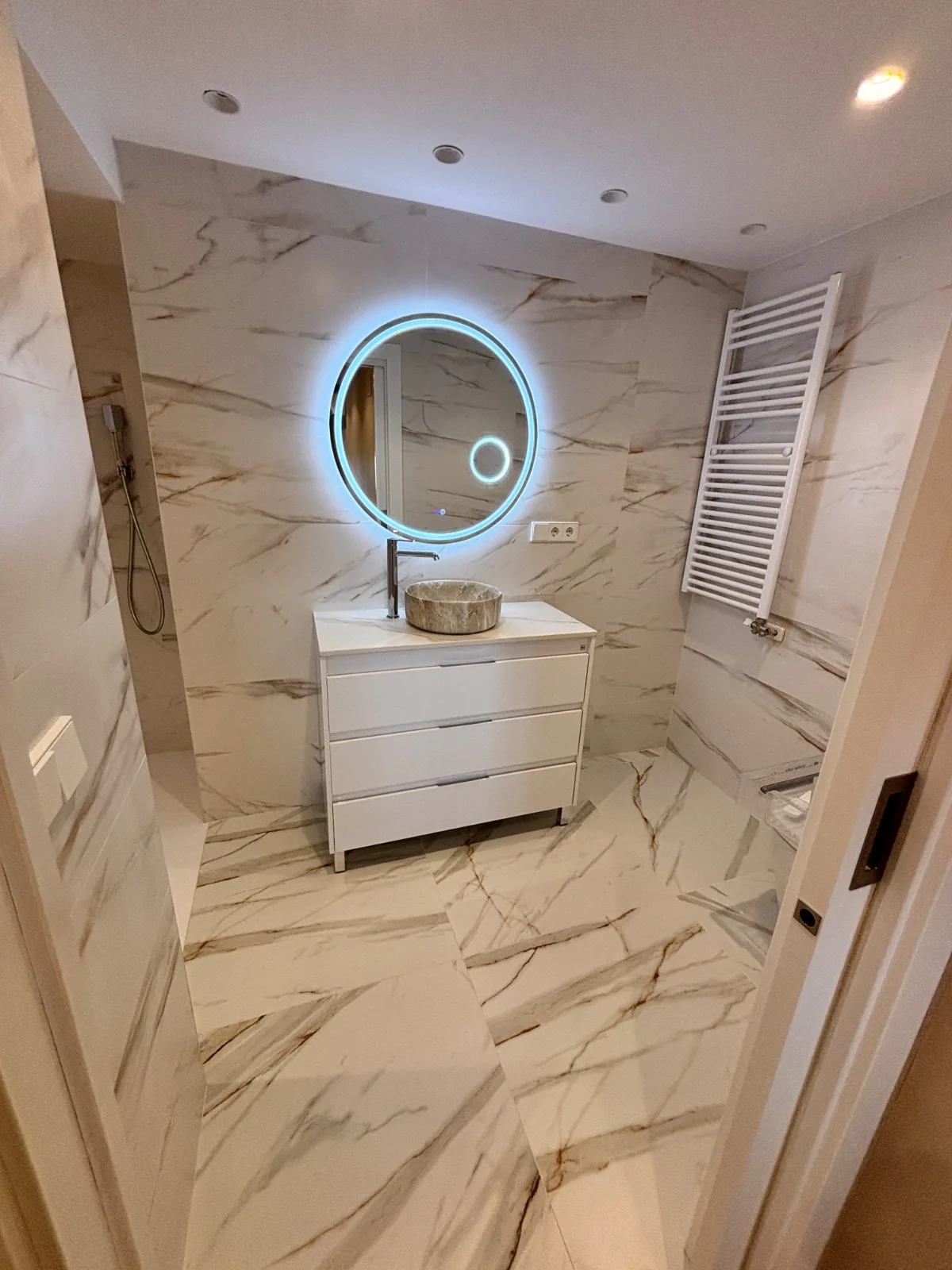 Baño reformado con mueble blanco, lavabo de piedra, espejo con luz led y radiador toallero
